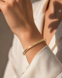 A close-up of a person's wrist wearing a slim gold bangle bracelet with small clear stones set along the band, paired with a white shirt in warm natural light.