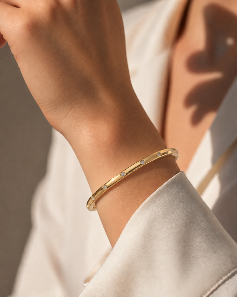 A close-up of a person's wrist wearing a slim gold bangle bracelet with small clear stones set along the band, paired with a white shirt in warm natural light.