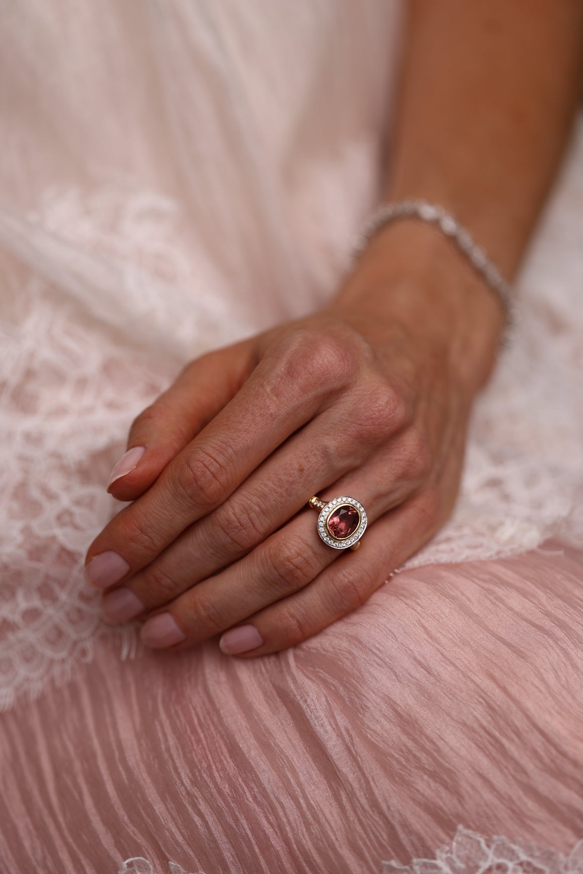 A close-up of a person's hand resting on soft pink lace fabric, wearing a gold ring with a round red gemstone surrounded by small clear stones, along with a silver-toned bracelet.