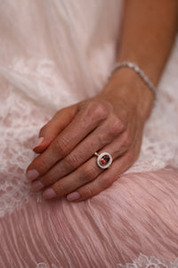 A close-up of a person's hand resting on soft pink lace fabric, wearing a gold ring with a round red gemstone surrounded by small clear stones, along with a silver-toned bracelet.