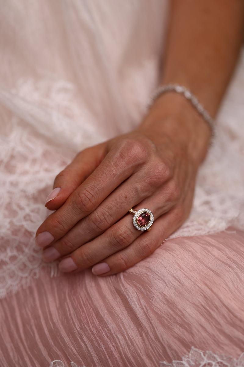 A close-up of a person's hand resting on soft pink lace fabric, wearing a gold ring with a round red gemstone surrounded by small clear stones, along with a silver-toned bracelet.