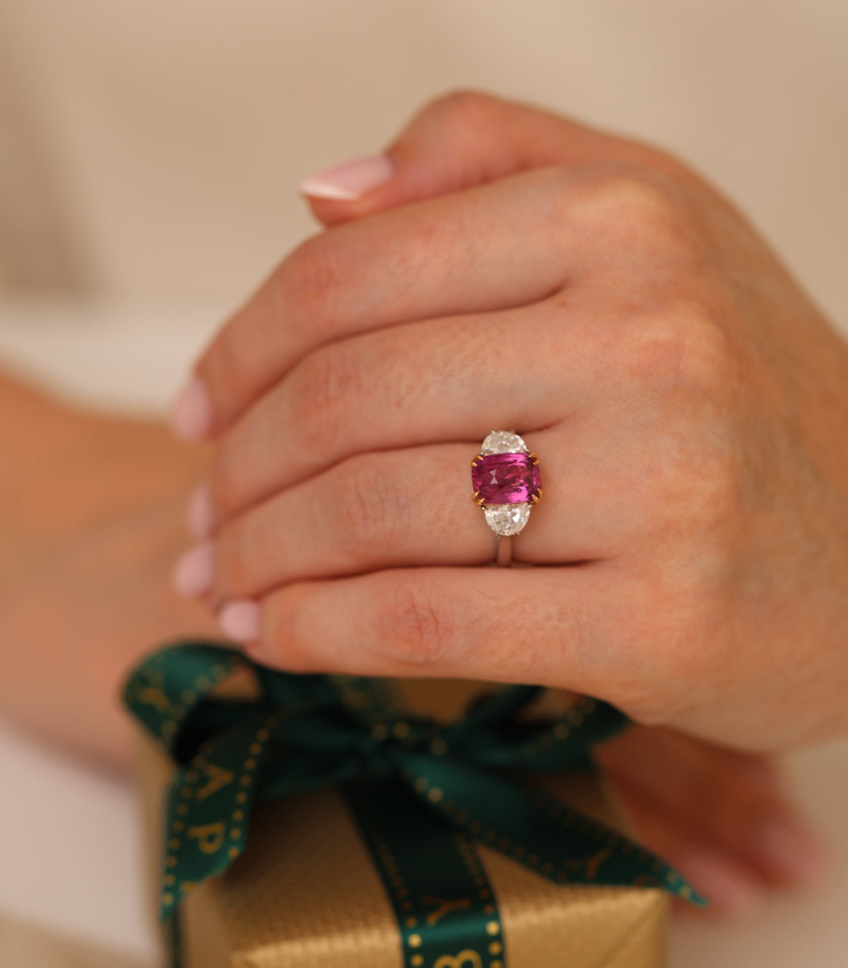 A close-up of a person's hand wearing a ring with a pink gemstone on a gold band, holding a gold gift box tied with a dark green ribbon.