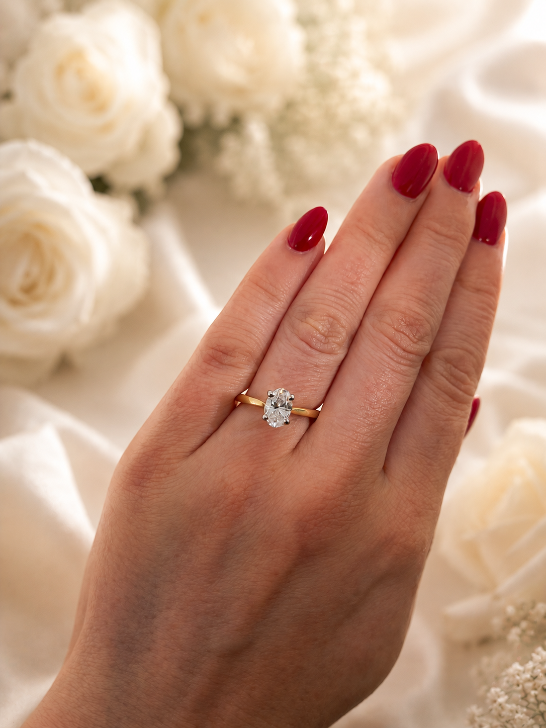 A gold engagement ring with a round-cut clear gemstone worn on a hand with red manicured nails, set against a soft background of white roses and draped fabric.