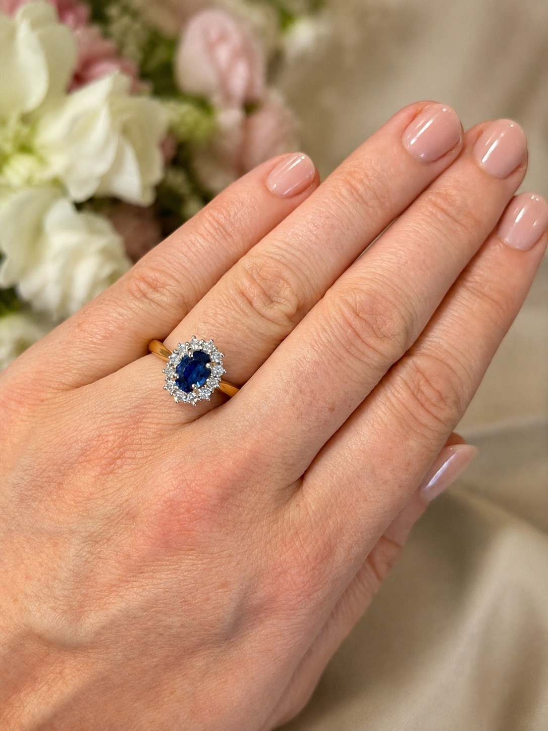 A close-up of a hand wearing a ring with a round blue center stone surrounded by small clear stones, set on a gold band, with a soft floral background.