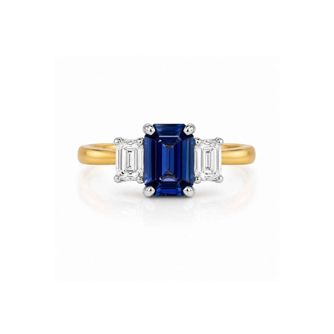A three-stone diamond ring featuring a central emerald-cut blue sapphire flanked by two baguette-cut diamonds, set in a yellow gold band with a white gold or platinum pavé setting.