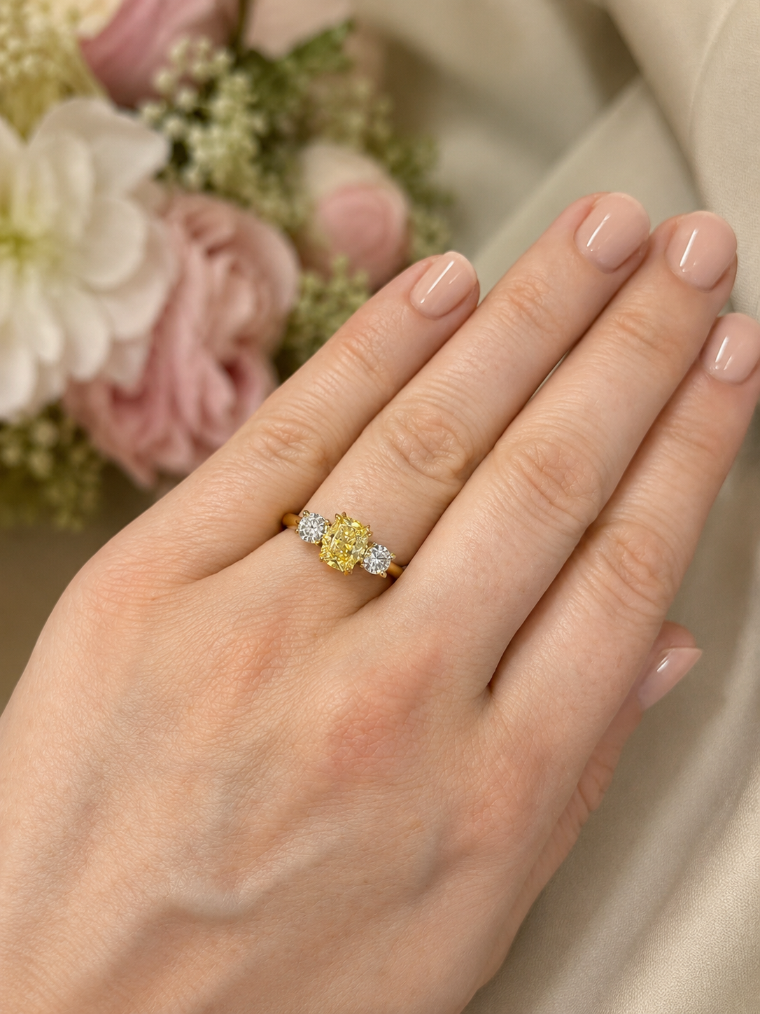 A close-up of a hand wearing a yellow diamond ring with small clear accent diamonds on a gold band, set against a soft beige fabric and blurred pink flowers.
