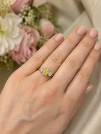 A close-up of a hand wearing a yellow diamond ring with small clear accent diamonds on a gold band, set against a soft beige fabric and blurred pink flowers.